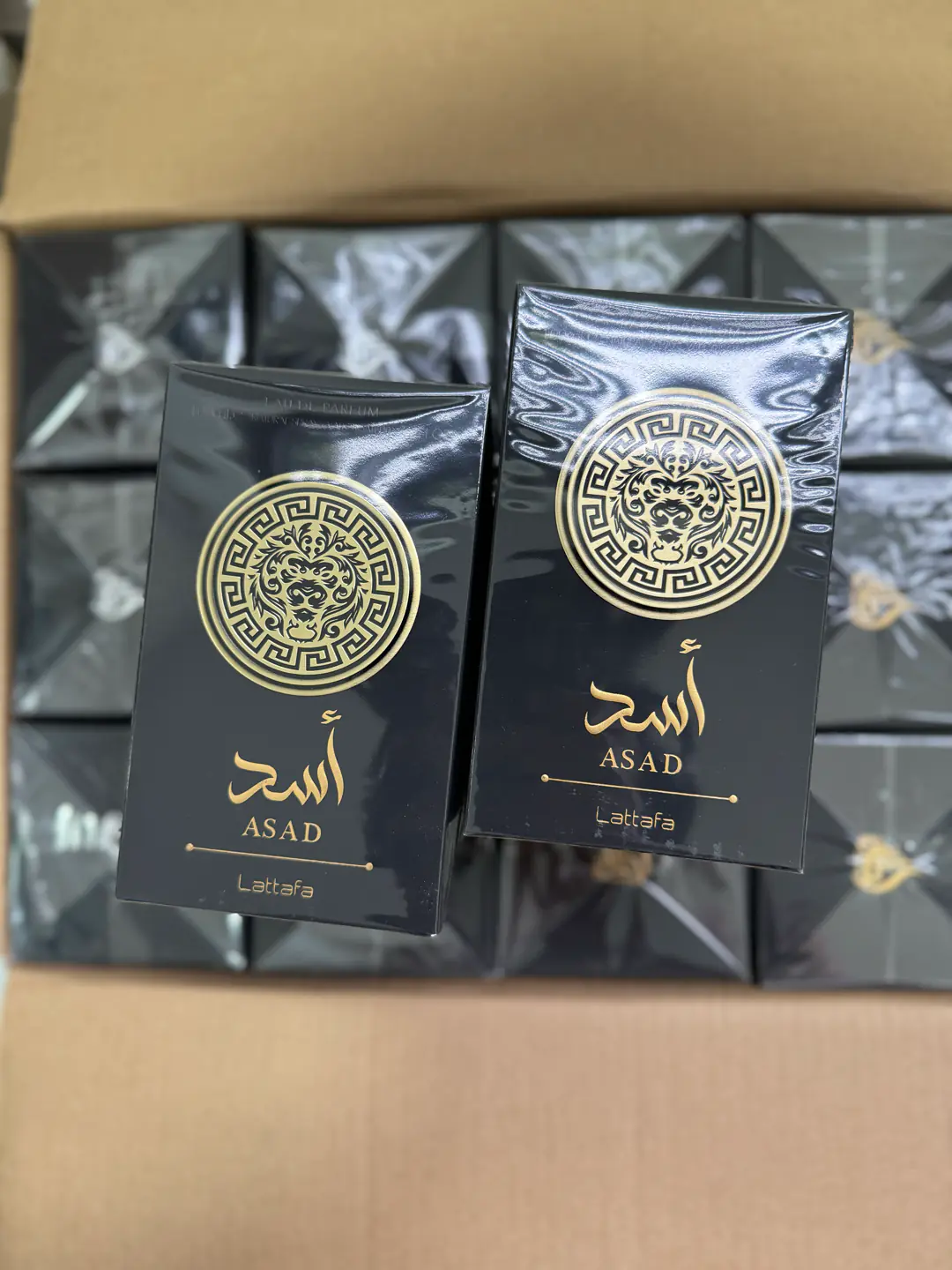 Dos cajas de Lattafa Asad selladas de fábrica sobre una caja abierta llena de perfumes Asad
